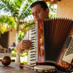 A musician playing a wooden accordion next to a güira and tambora on a rustic patio.