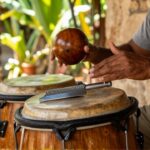Hands play a metal güira and a leather conga drum in a sunlit tropical courtyard.