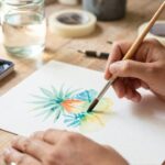 Hands use a squirrel hair brush to mix cerulean and ochre pigments on a ceramic palette.