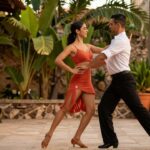 A female dancer performs energetic Merengue steps next to a male dancer doing a slow Bachata sway in a sun-drenched courtyard.