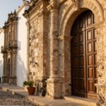 A weathered limestone archway and dark wooden door stand on a sun-drenched cobblestone street.