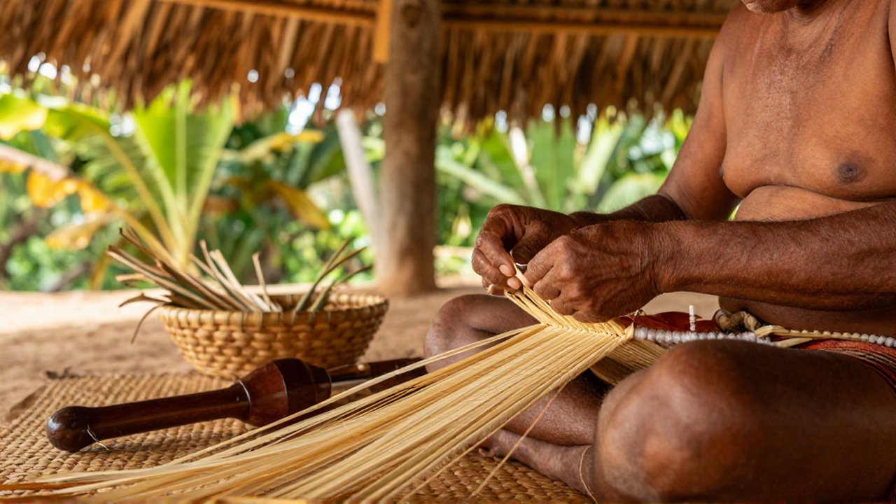 An artisan weaves dried palm fibers and cotton threads on a mat inside a thatched shelter.